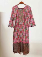 A woman modeling a multicolor premium cotton all-over printed stitched three-piece dress, featuring a geometric box pattern and traditional paisley hem, paired with a matching zigzag printed chiffon dupatta and black printed trousers.